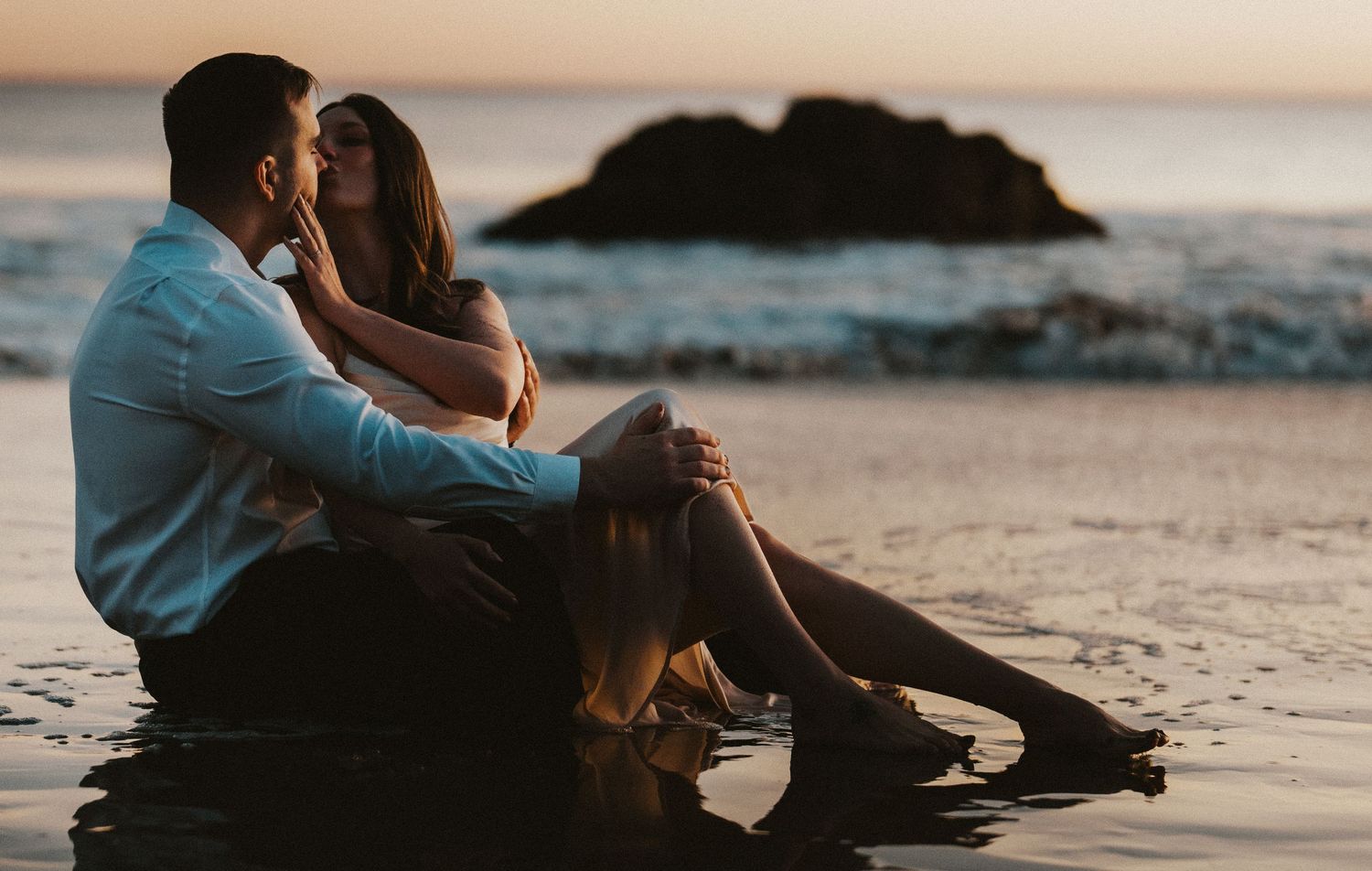 Romantic couple sitting together on a wet beach shore during a stunning golden sunset.