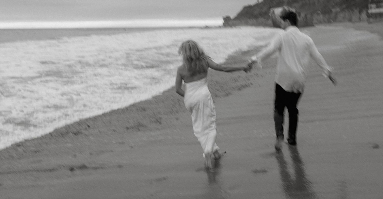 Black and white photo of couple holding hands while walking along shoreline on a sandy beach.