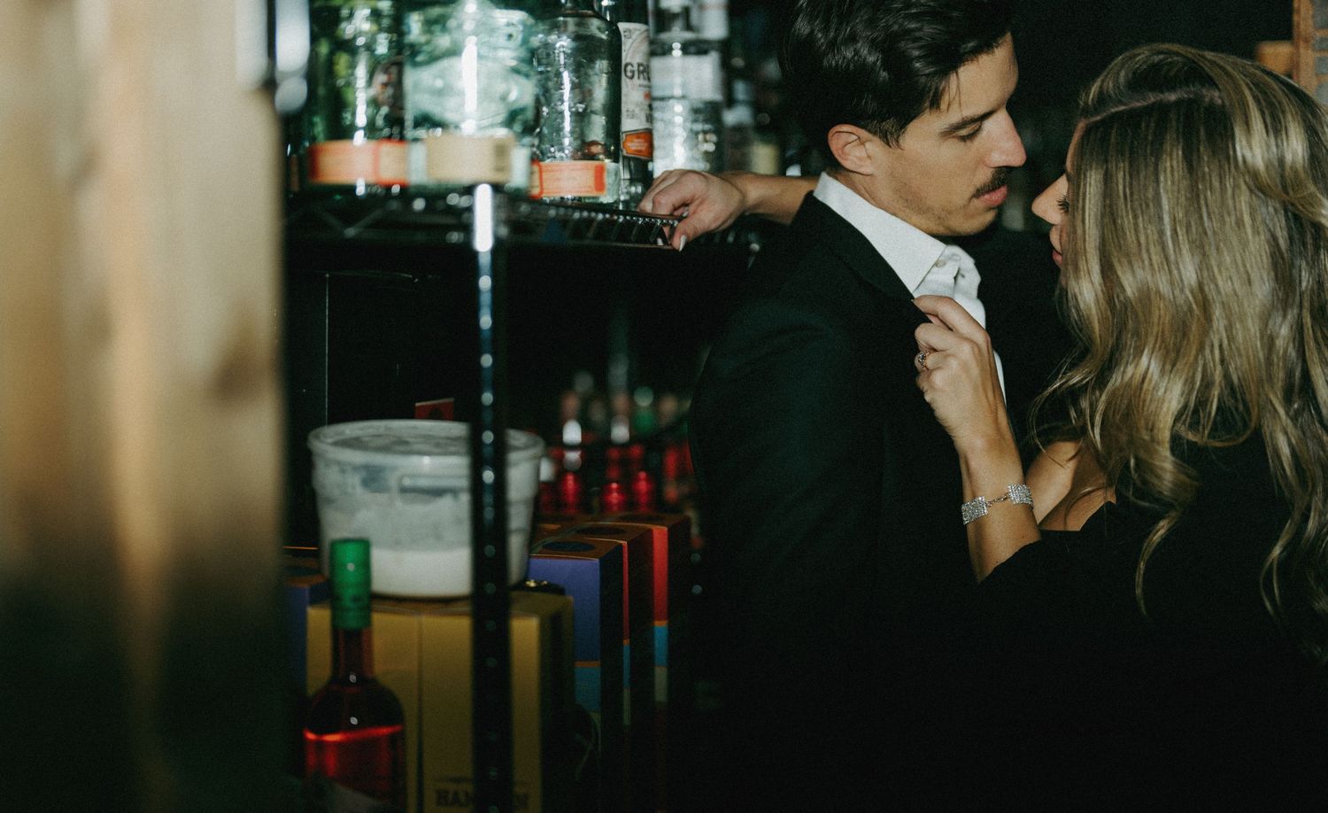 Couple sharing an intimate moment at a bar, woman in black dress adjusting man's tie.