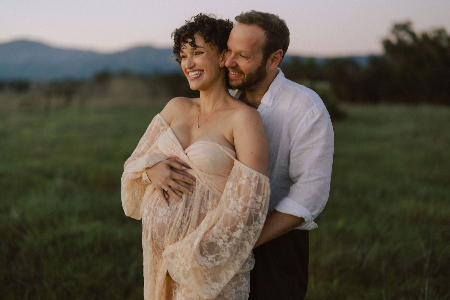 Couple embracing in green meadow at dusk, woman in elegant lace maternity gown.