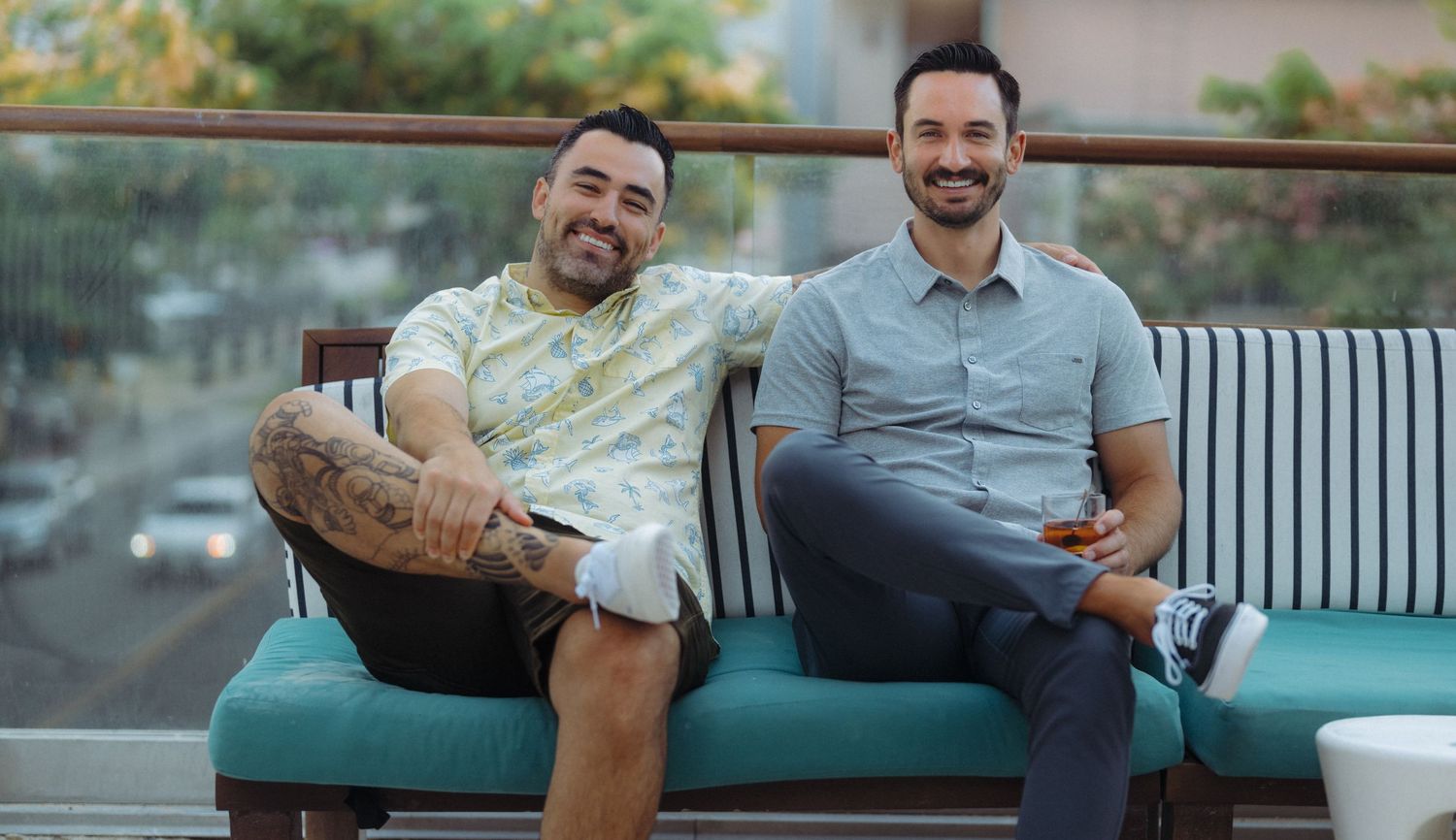 Two smiling men sitting together on a teal couch on an outdoor balcony in casual summer attire.