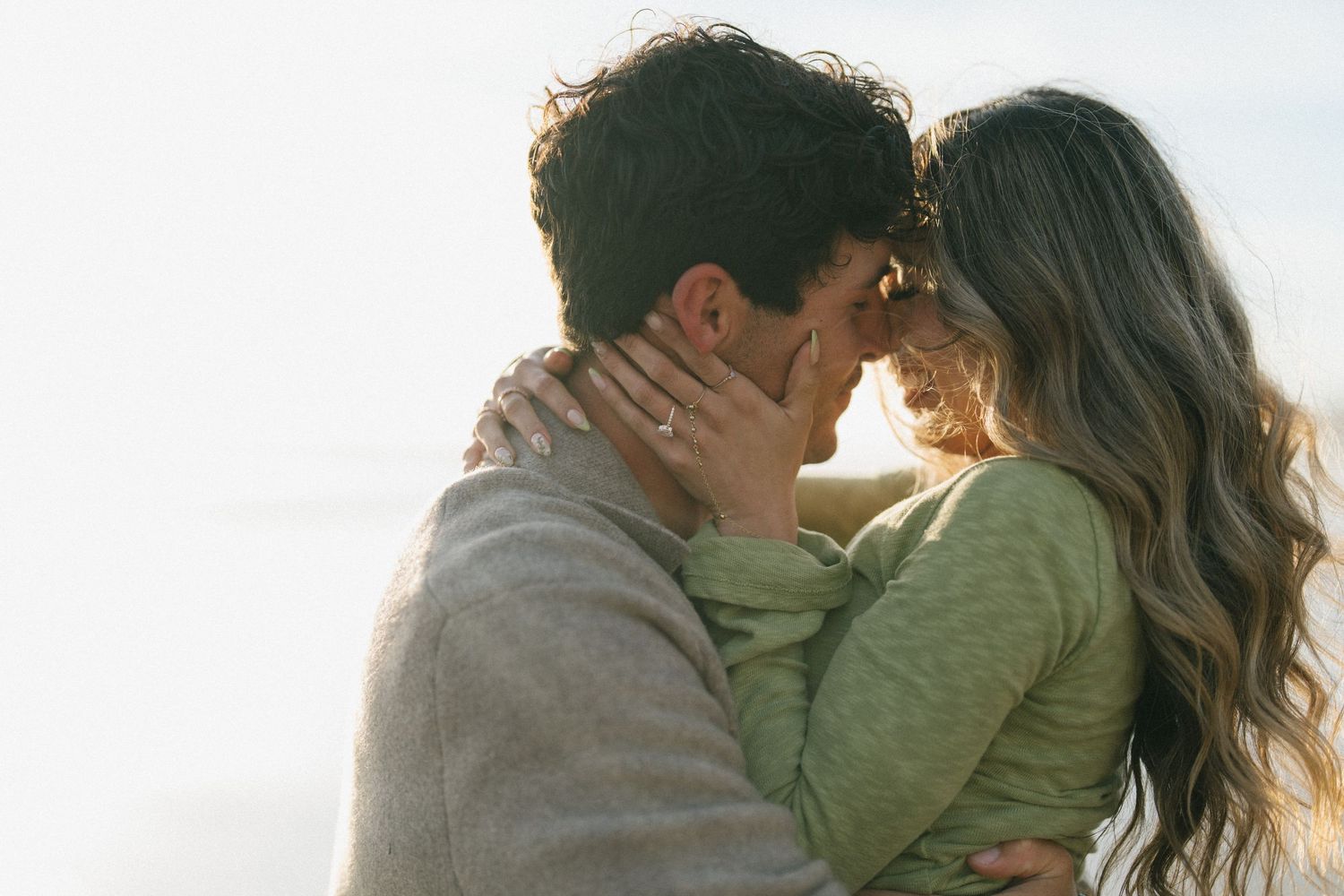 Couple sharing a tender kiss outdoors in soft golden backlight, woman wearing engagement ring.