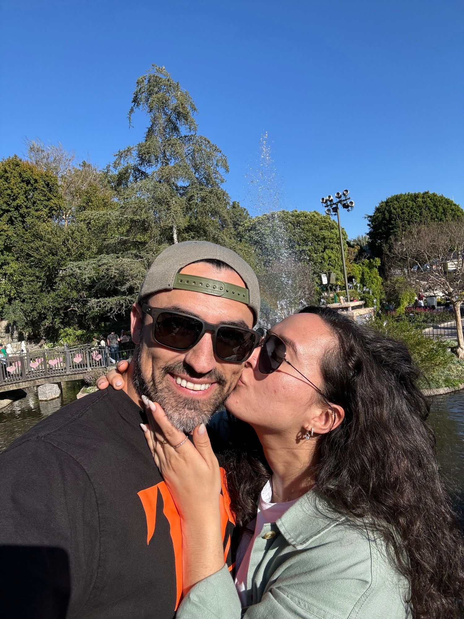 Couple smiling and sharing a kiss outdoors near fountain on sunny clear day.