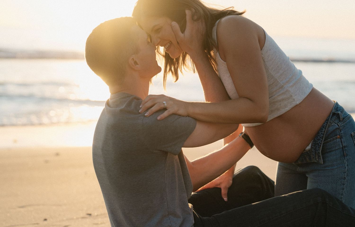 Couple sharing romantic moment on beach at sunset, pregnant woman leaning in for kiss.
