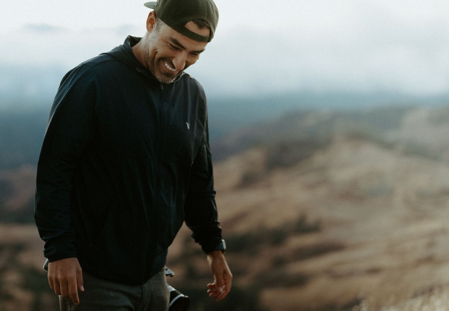 Man in black jacket wearing backward cap looking down on a misty mountain hiking trail.