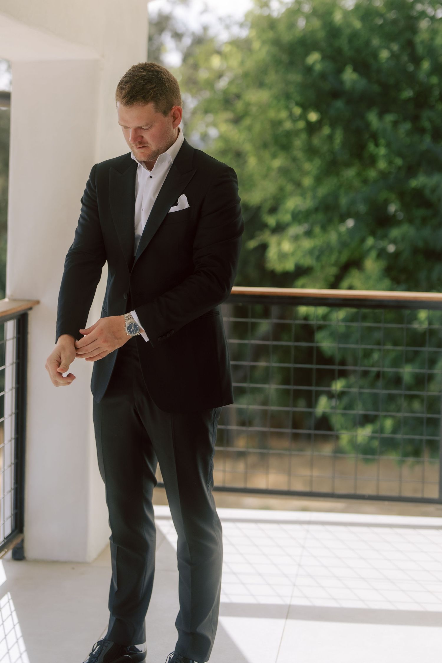 Dapper groom in black tuxedo adjusting cufflinks on outdoor balcony surrounded by lush green trees.