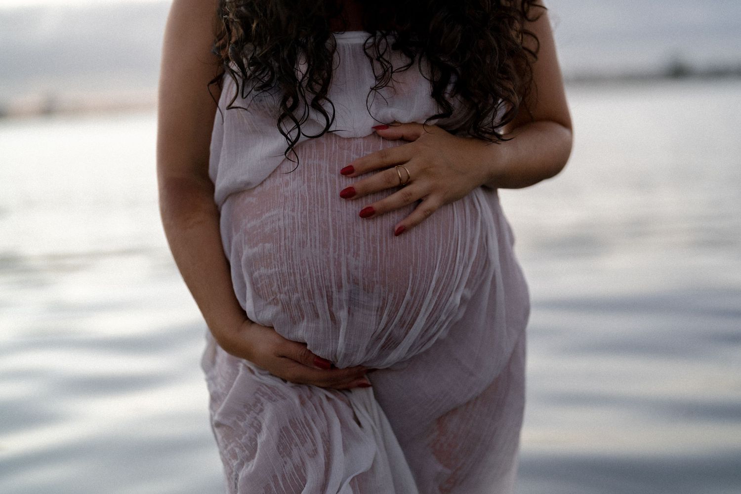 Pregnant woman in sheer white dress cradles baby bump near water during maternity session.