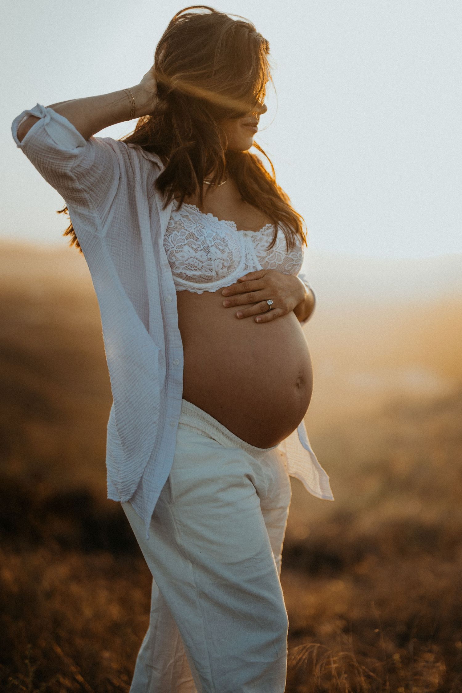 Pregnant woman in white lace bralette and pants holding baby bump during beautiful golden hour sunset.