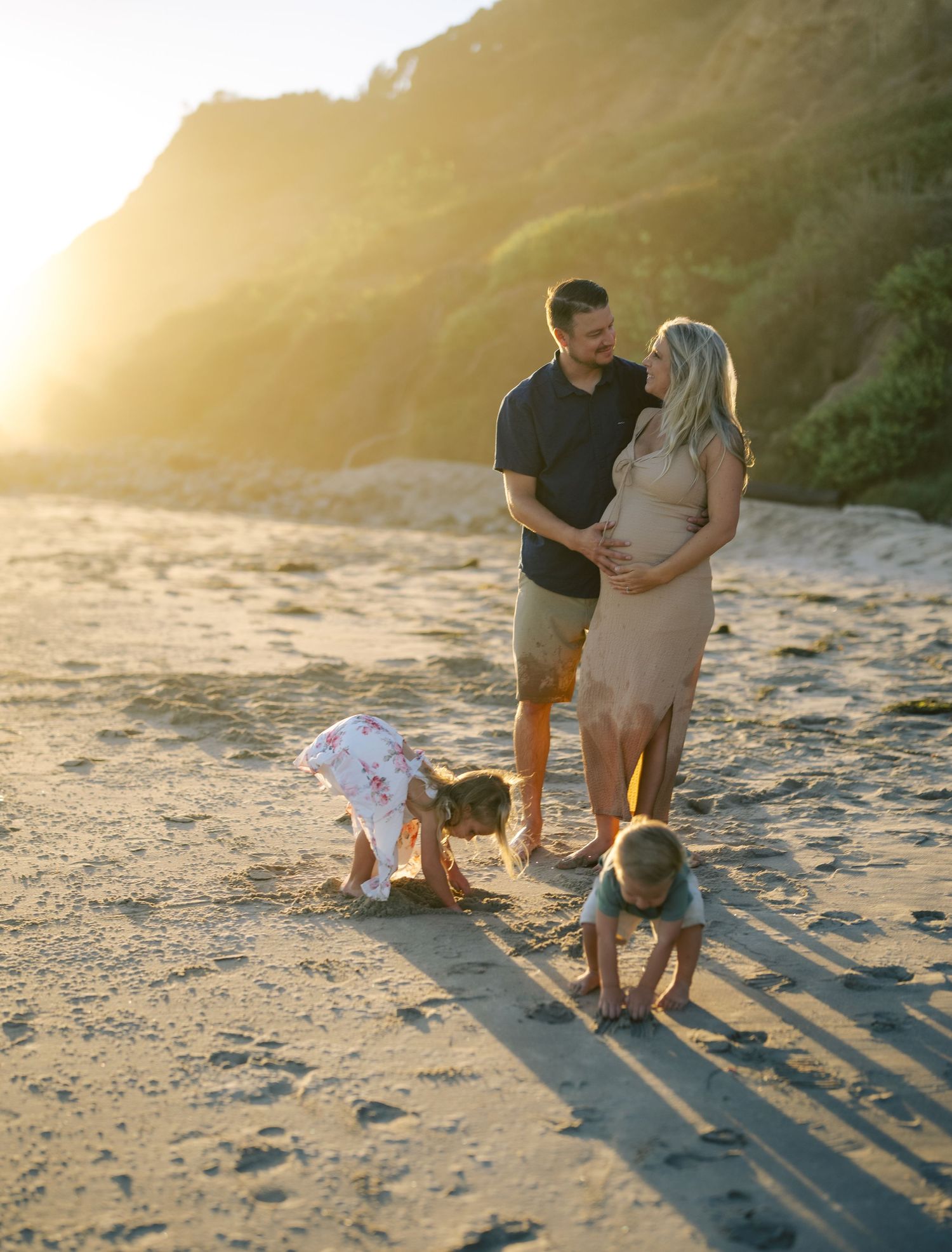 Pregnant family with two young children playing on sandy beach during golden hour sunset.