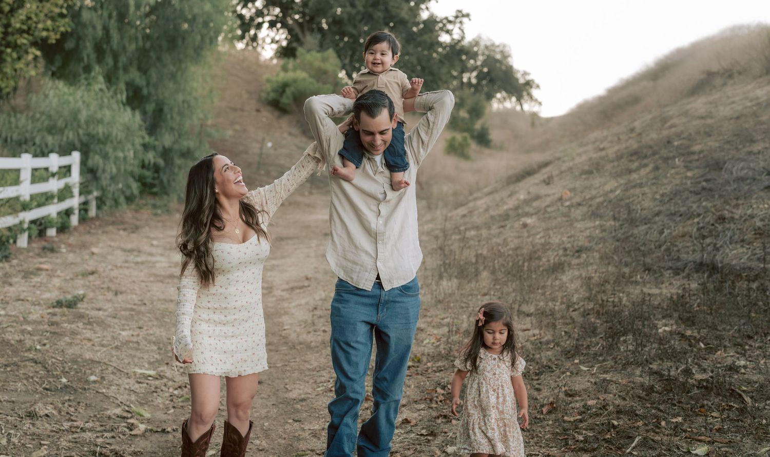 Happy family of four walking along a sunny hillside trail with a white fence backdrop.