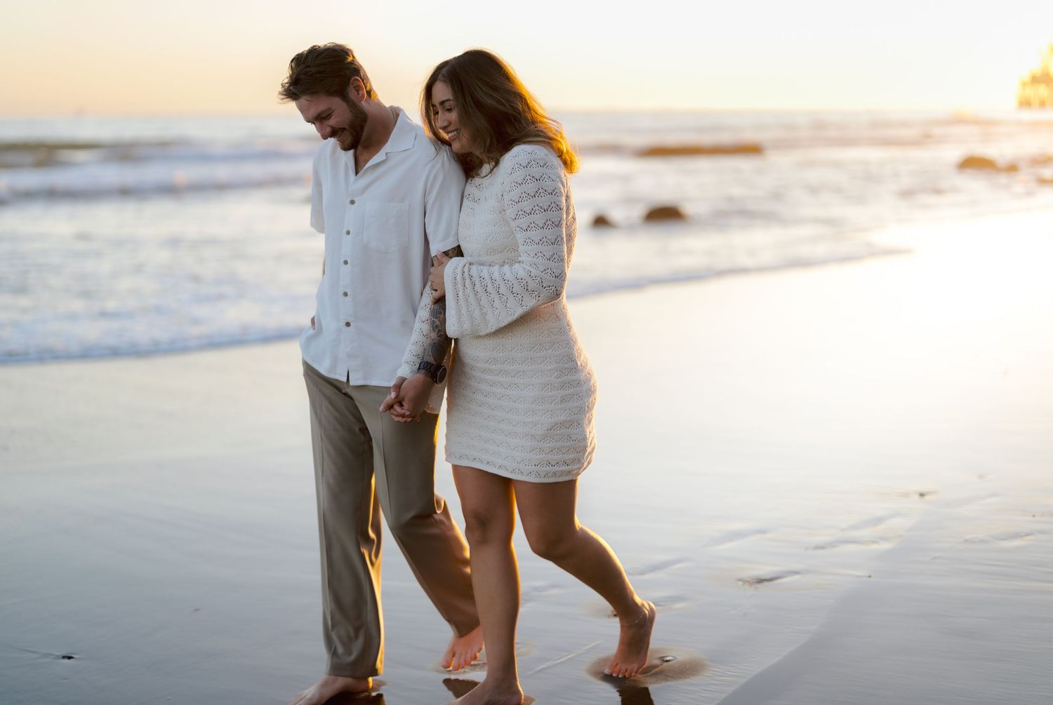 Romantic couple walking hand in hand along sandy beach at golden hour sunset.