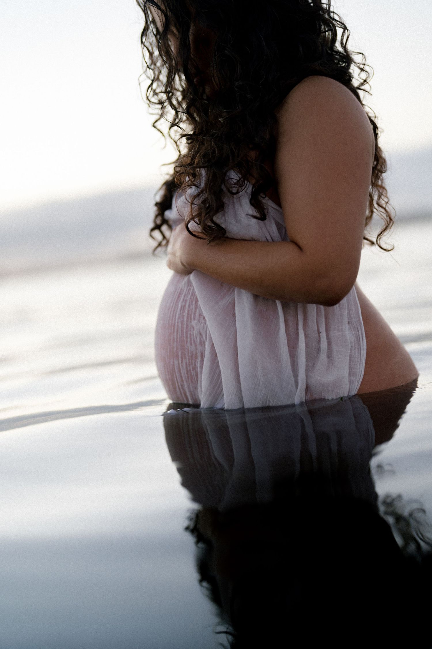 Pregnant woman in sheer white dress stands in shallow water cradling her baby bump.