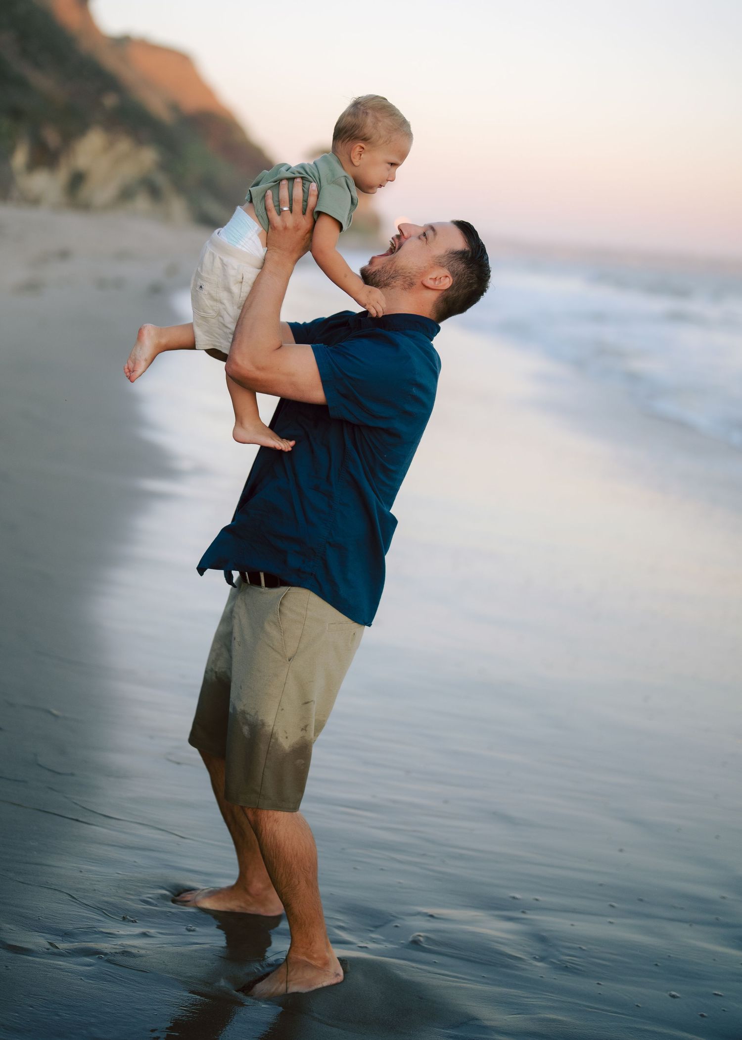 Father joyfully lifting smiling baby in the air on a serene beach at sunset.