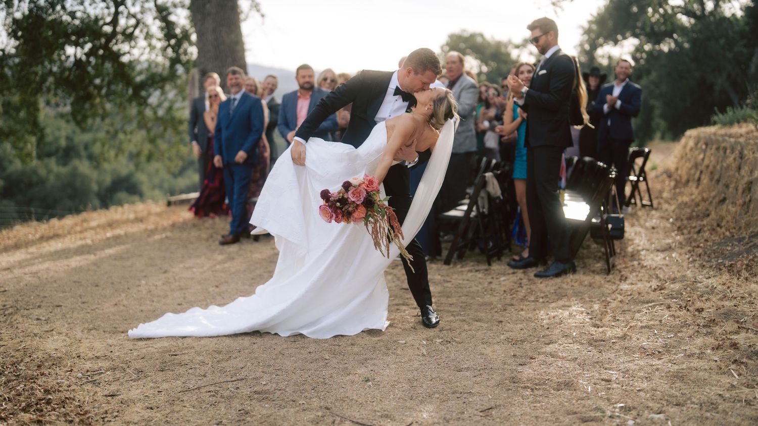 Groom dips bride in romantic kiss after outdoor wedding ceremony with guests applauding in background.
