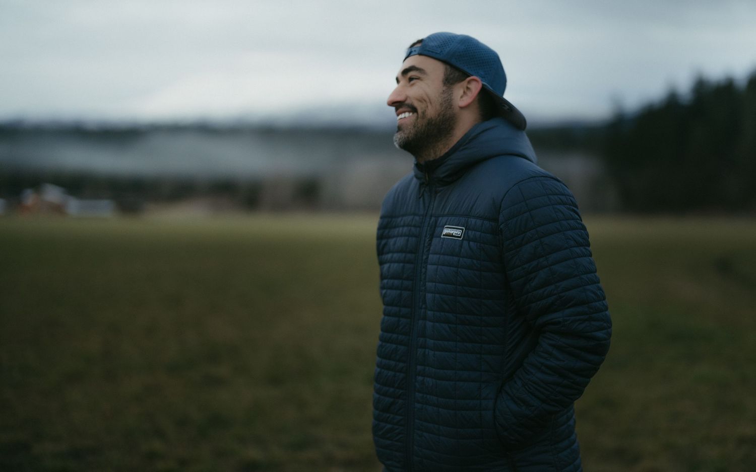 Smiling bearded man in navy Patagonia puffer jacket and beanie standing in an open field on a cloudy day.