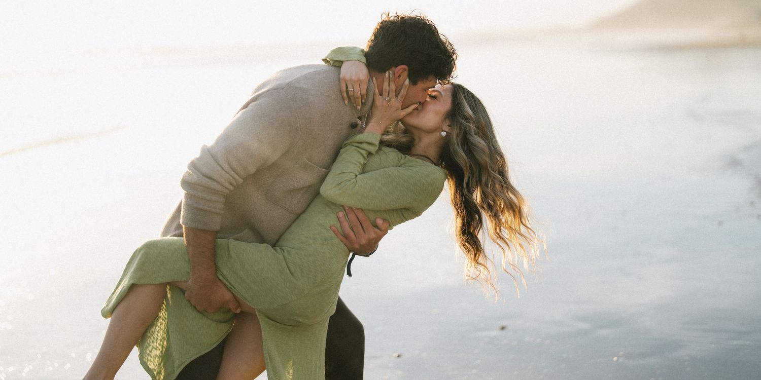Couple sharing a romantic kiss on a sunlit beach, woman in green dress lifted by partner.