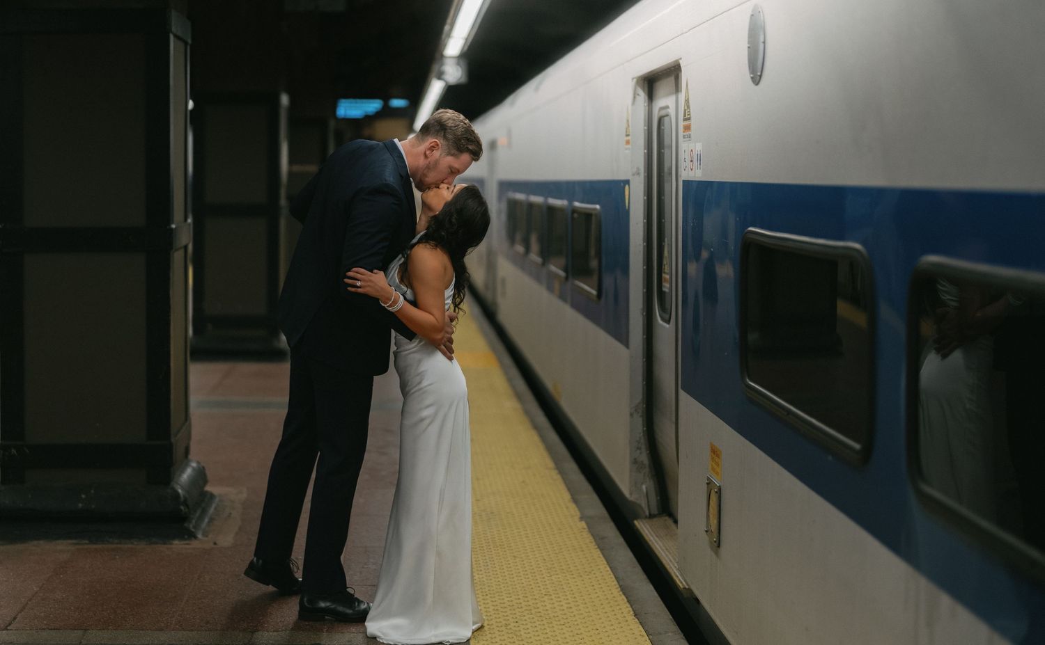 Couple sharing a romantic kiss on a train platform, dressed in elegant wedding attire.