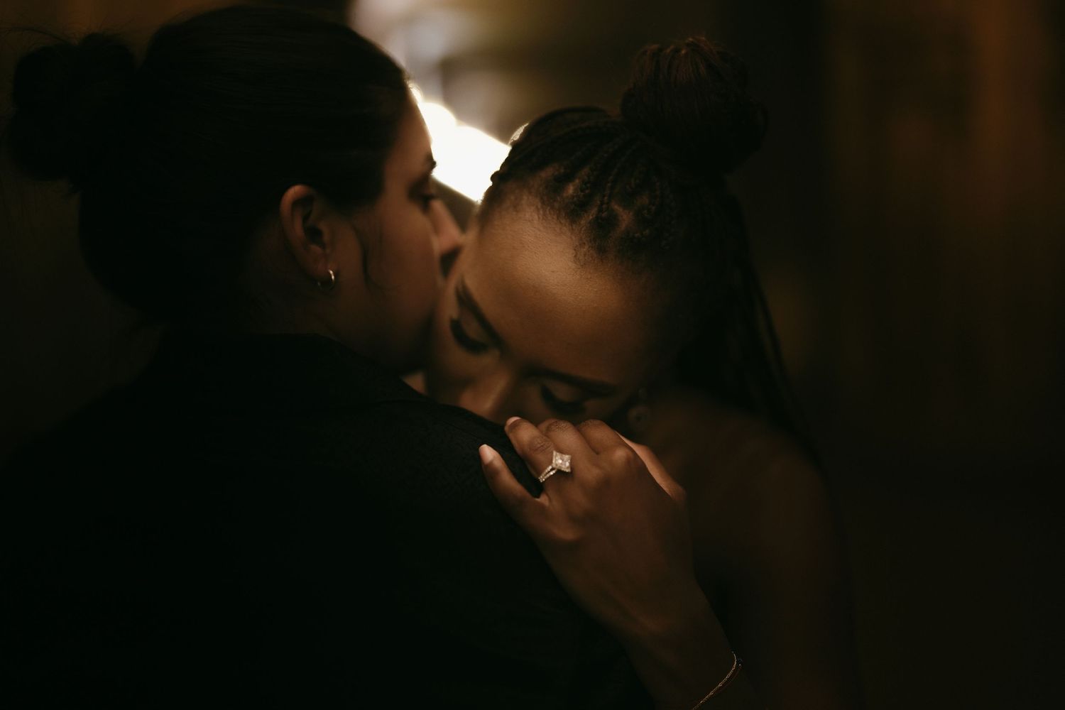 Two women share an intimate moment in dim romantic lighting, one tenderly kissing the other.