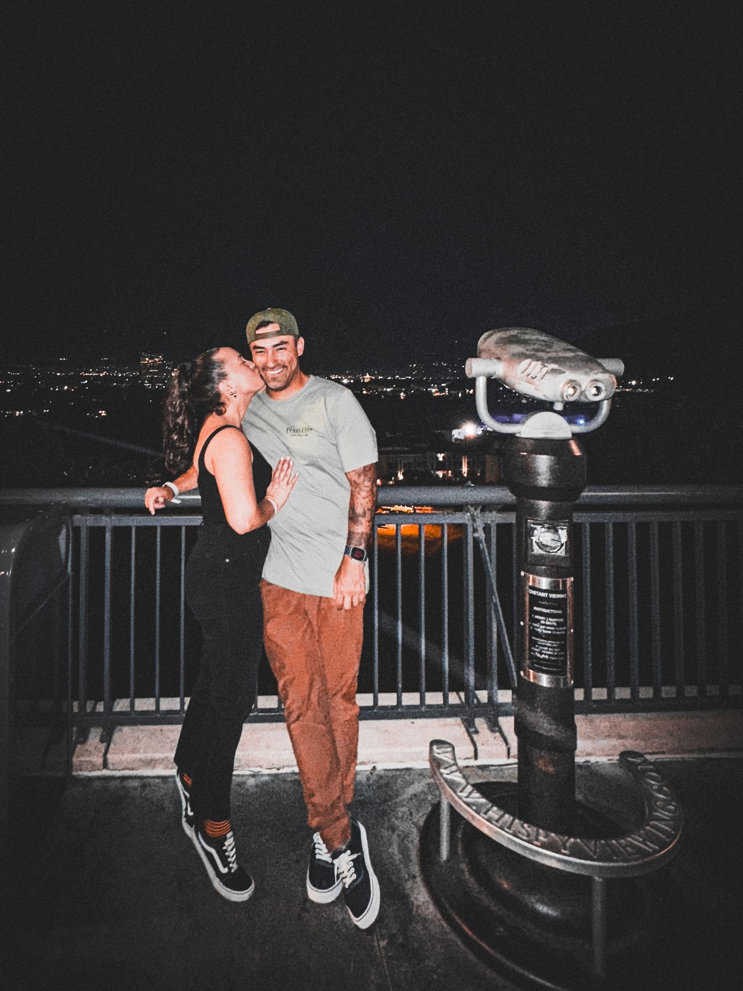 Couple embracing at night overlook with city lights and coin-operated binoculars nearby.