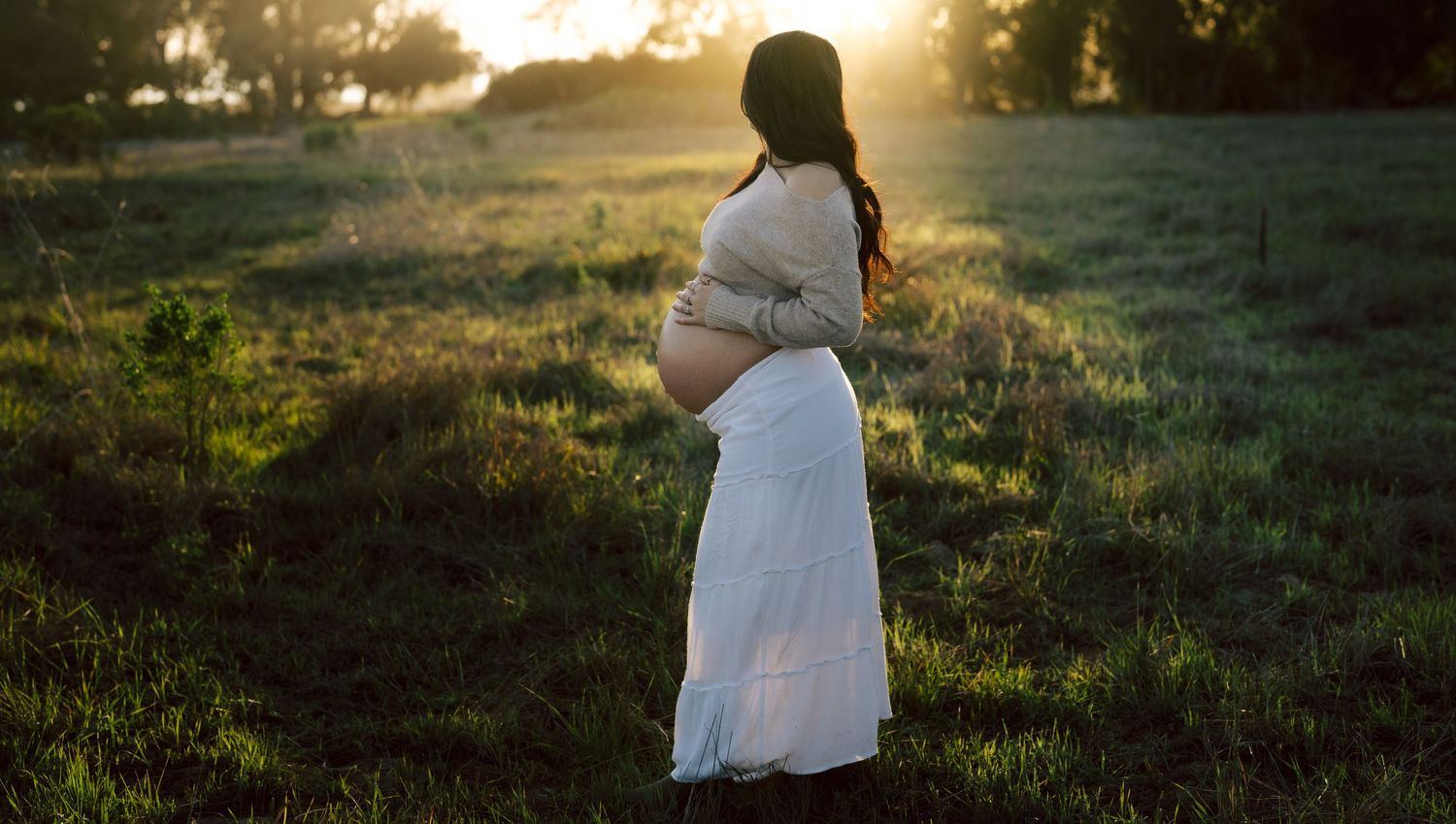 Pregnant woman in white dress standing alone in field during golden sunset.