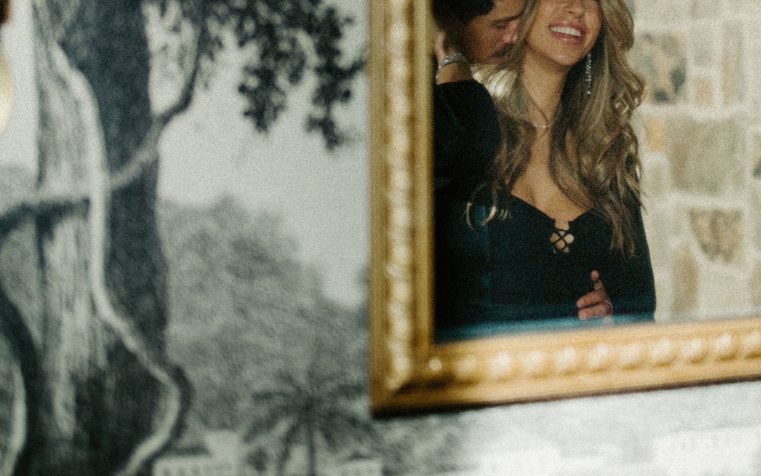 Couple smiling and embracing reflected in a gold-framed mirror with scenic mural wallpaper backdrop.