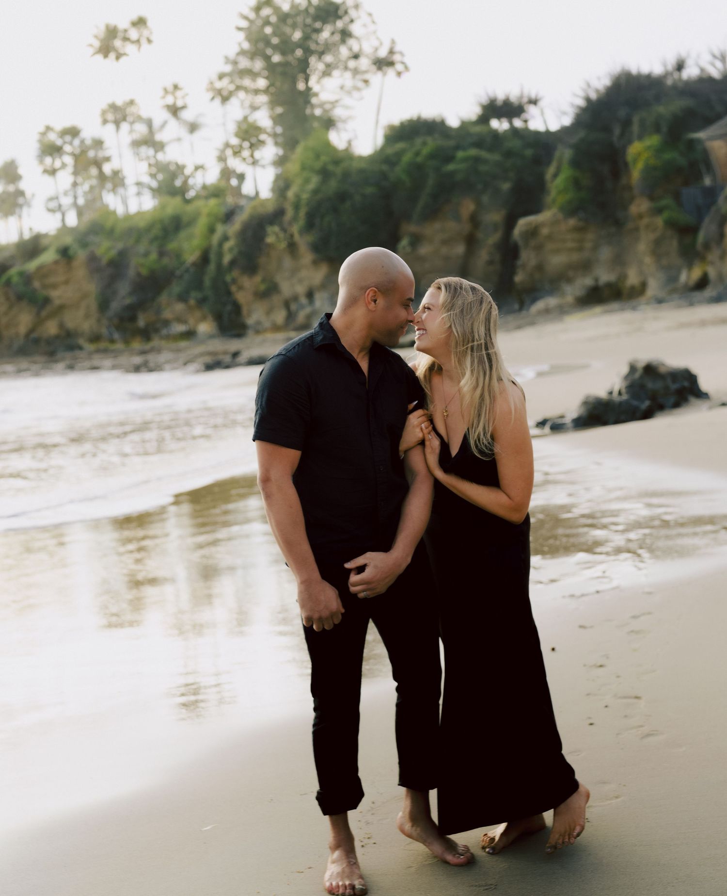 Couple in matching black outfits sharing romantic moment on reflective wet sand beach.