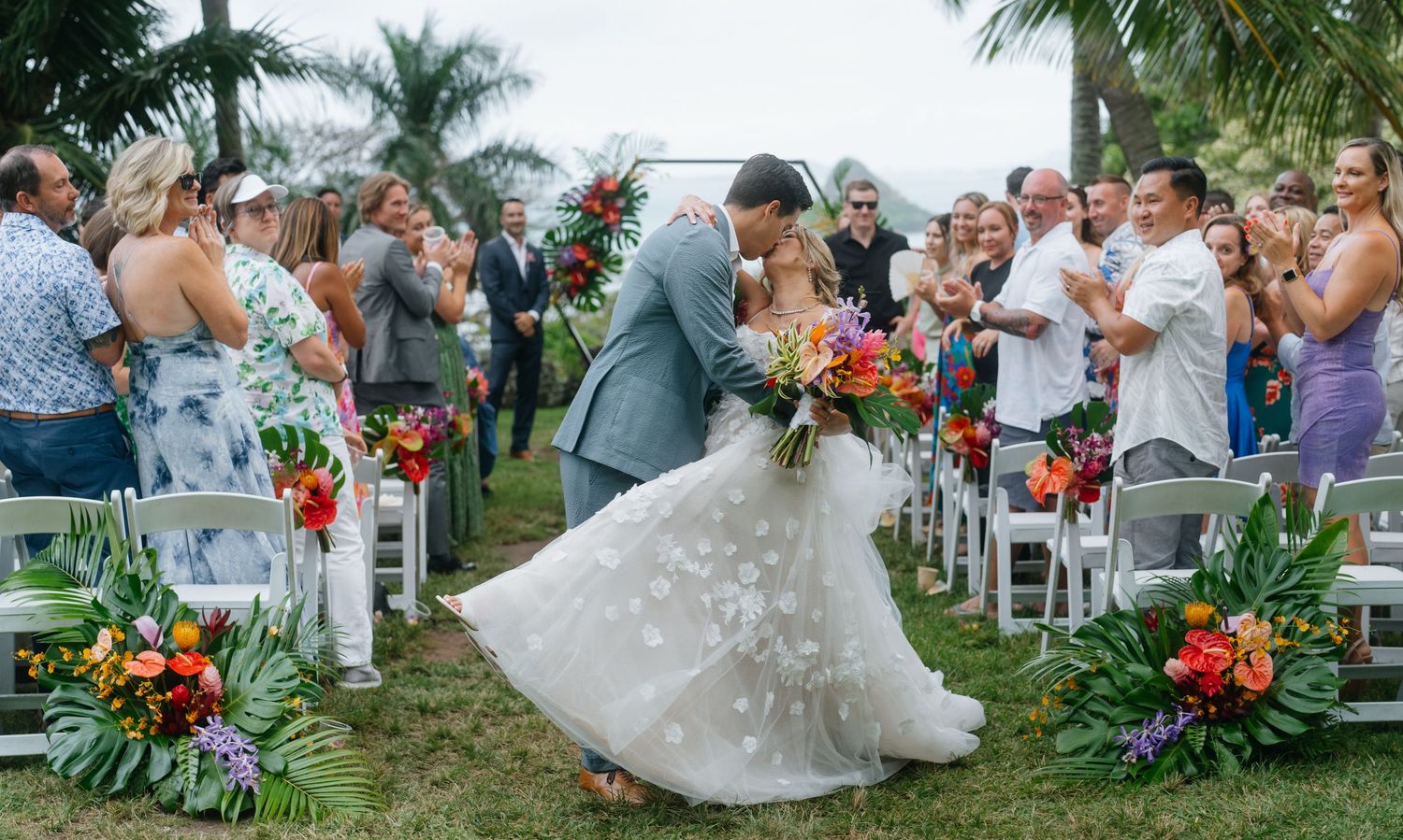 Bride and groom sharing romantic kiss in aisle after outdoor tropical wedding ceremony with colorful floral arrangements.