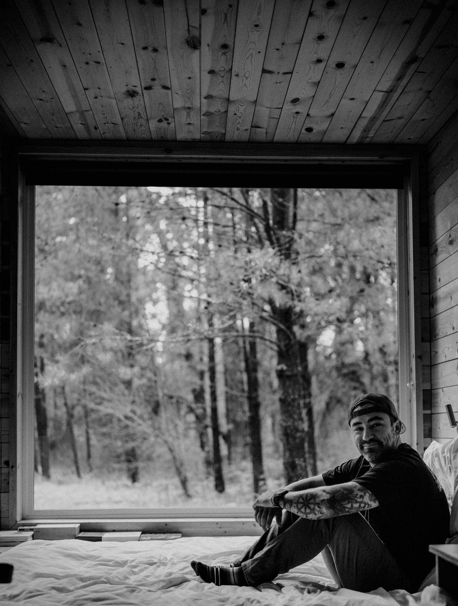 Black and white photo of tattooed man sitting on bed in rustic cabin with large forest window behind him.
