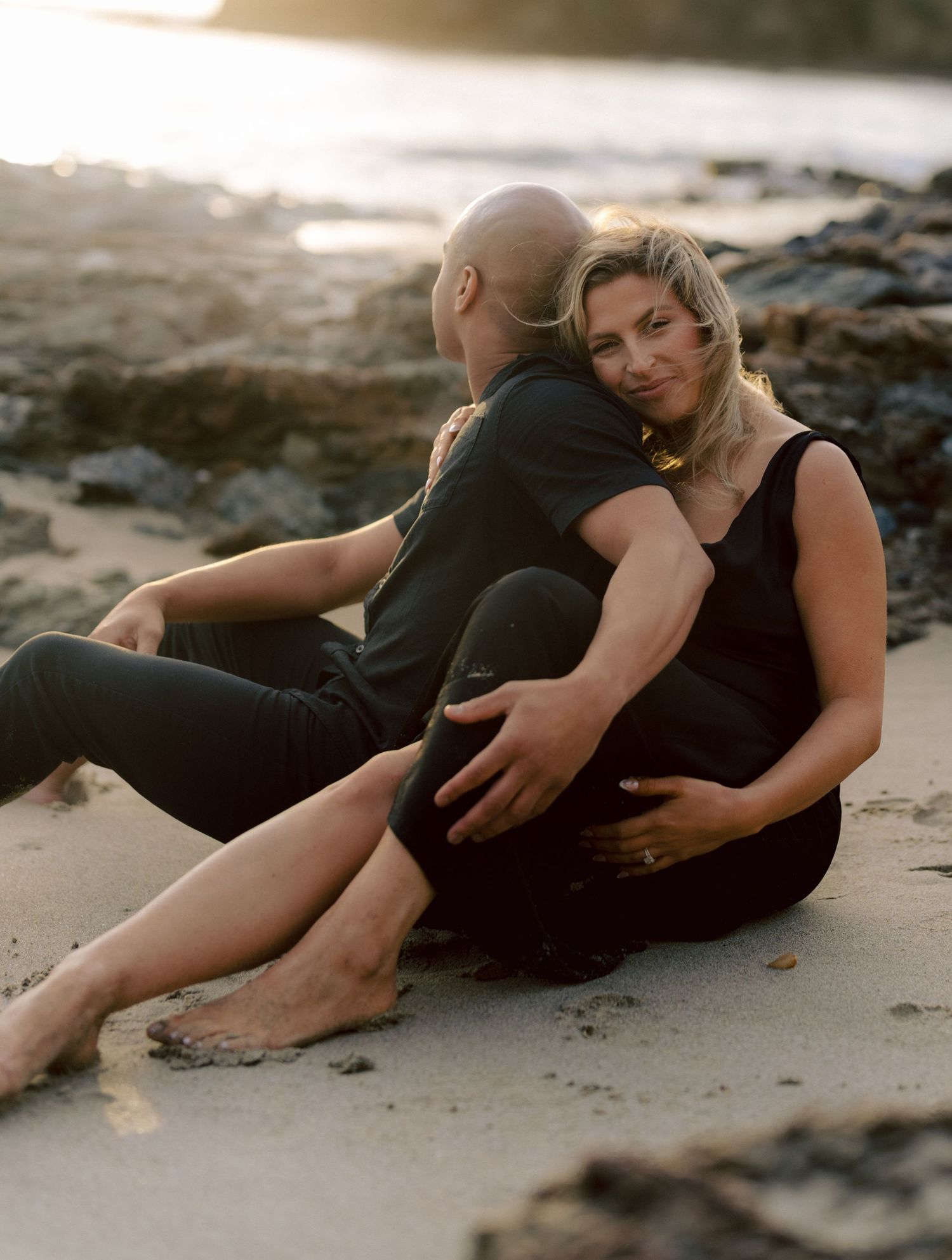 Couple sitting on beach sand at golden hour, woman leaning against man in matching black outfits.