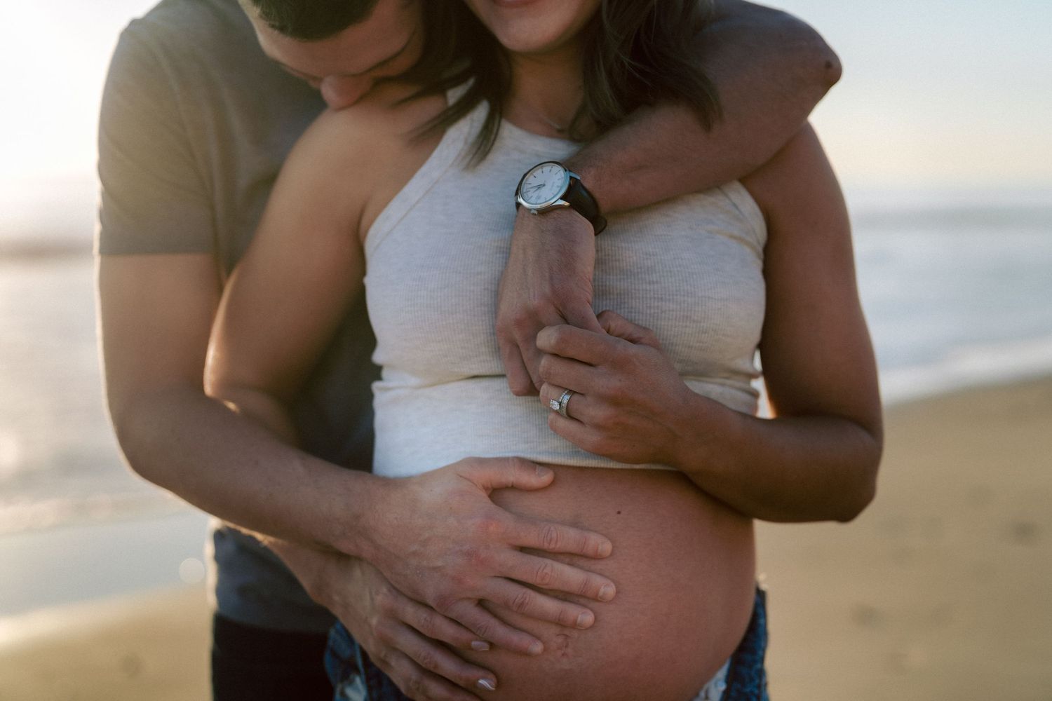 Couple holding pregnant belly together on beach at sunset, wearing casual clothes.