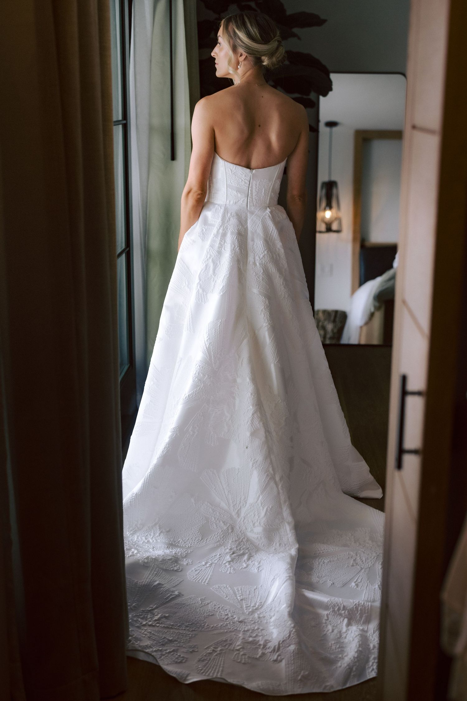 Bride in stunning strapless white ball gown wedding dress viewed from behind near doorway with mirror reflection.
