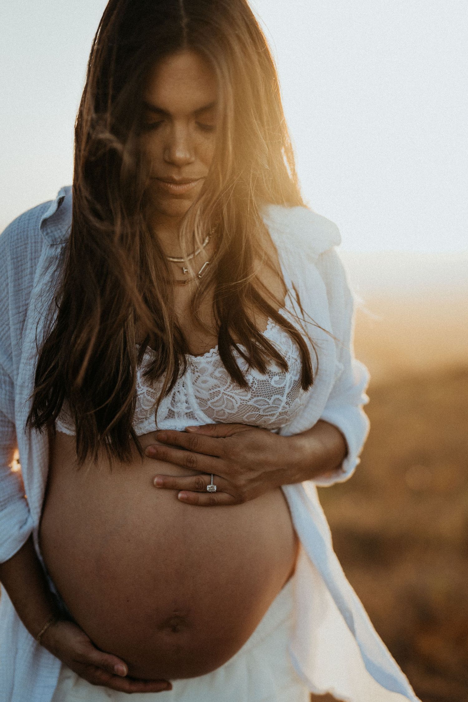 Pregnant woman in white lace bralette and open shirt cradling her baby bump at golden sunset.