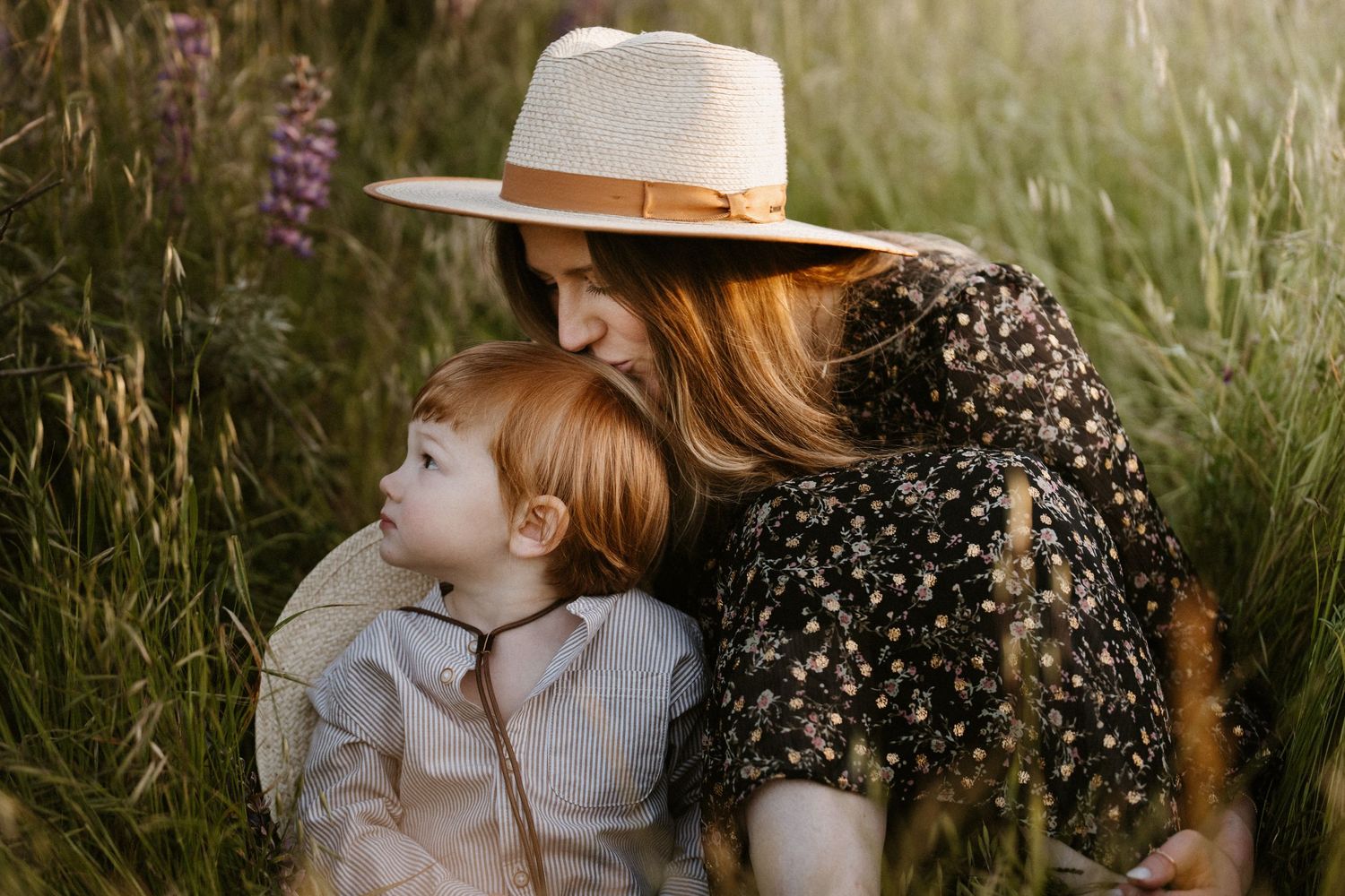 Tender moment of a mother in a straw hat embracing her red-haired toddler in tall grass.