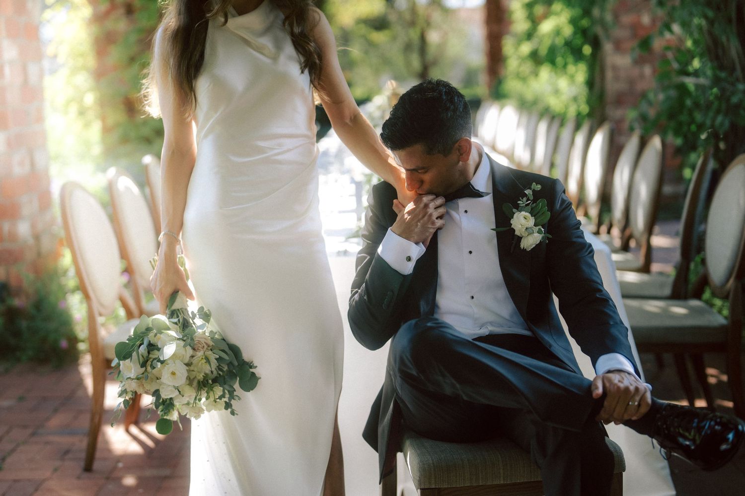 Groom in black tuxedo kisses bride's hand while seated as she holds white floral bouquet.