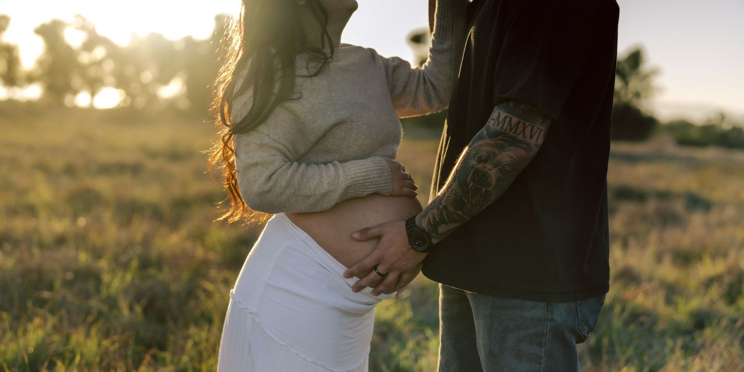 Couple embracing outdoors at golden hour, hands cradling pregnant belly in sunlit field.