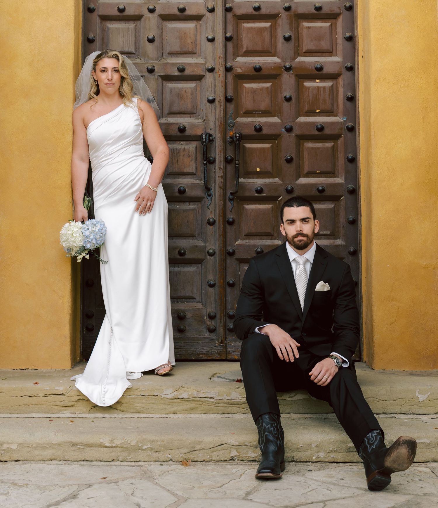Bride in white one-shoulder gown and groom in black suit pose before ornate wooden doors.
