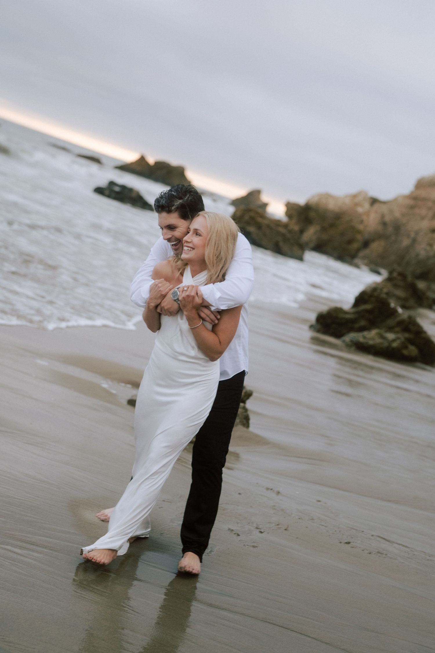Happy couple embracing and dancing on wet sandy beach at dusk with rocky coastline in background.