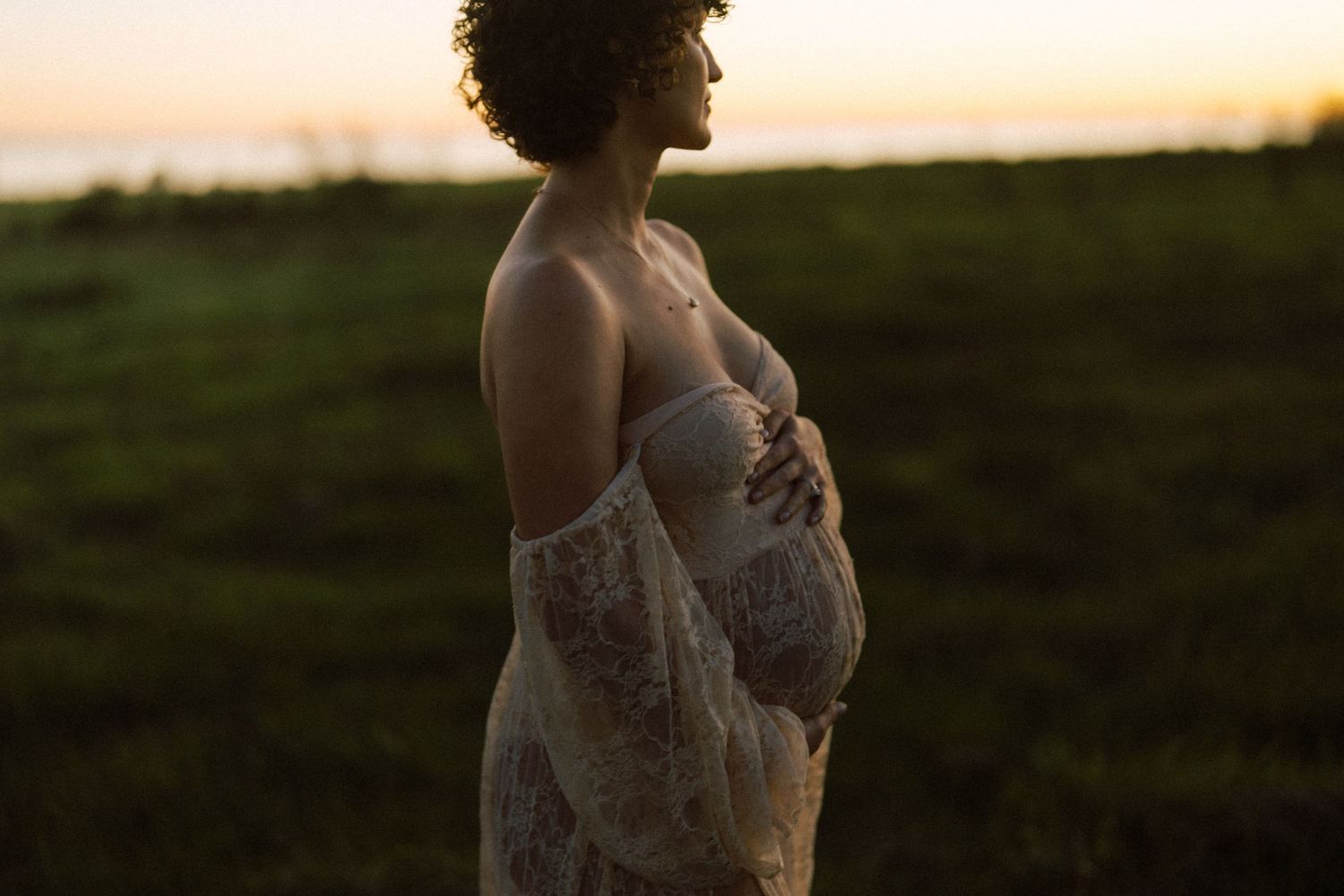 Pregnant woman in sheer lace dress holding belly during golden sunset outdoors.