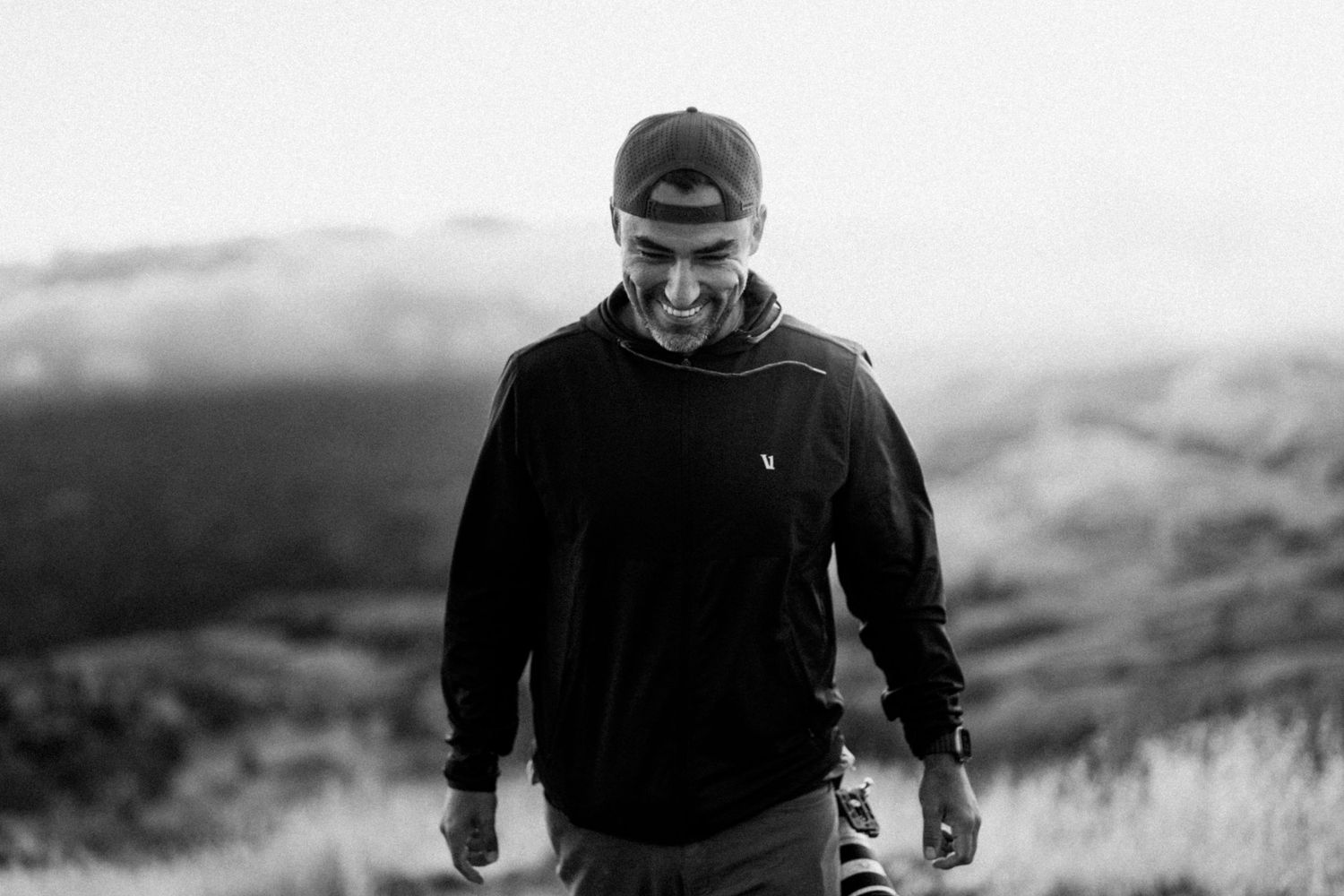 Man in black hoodie and cap smiling outdoors with misty mountain landscape in black and white.