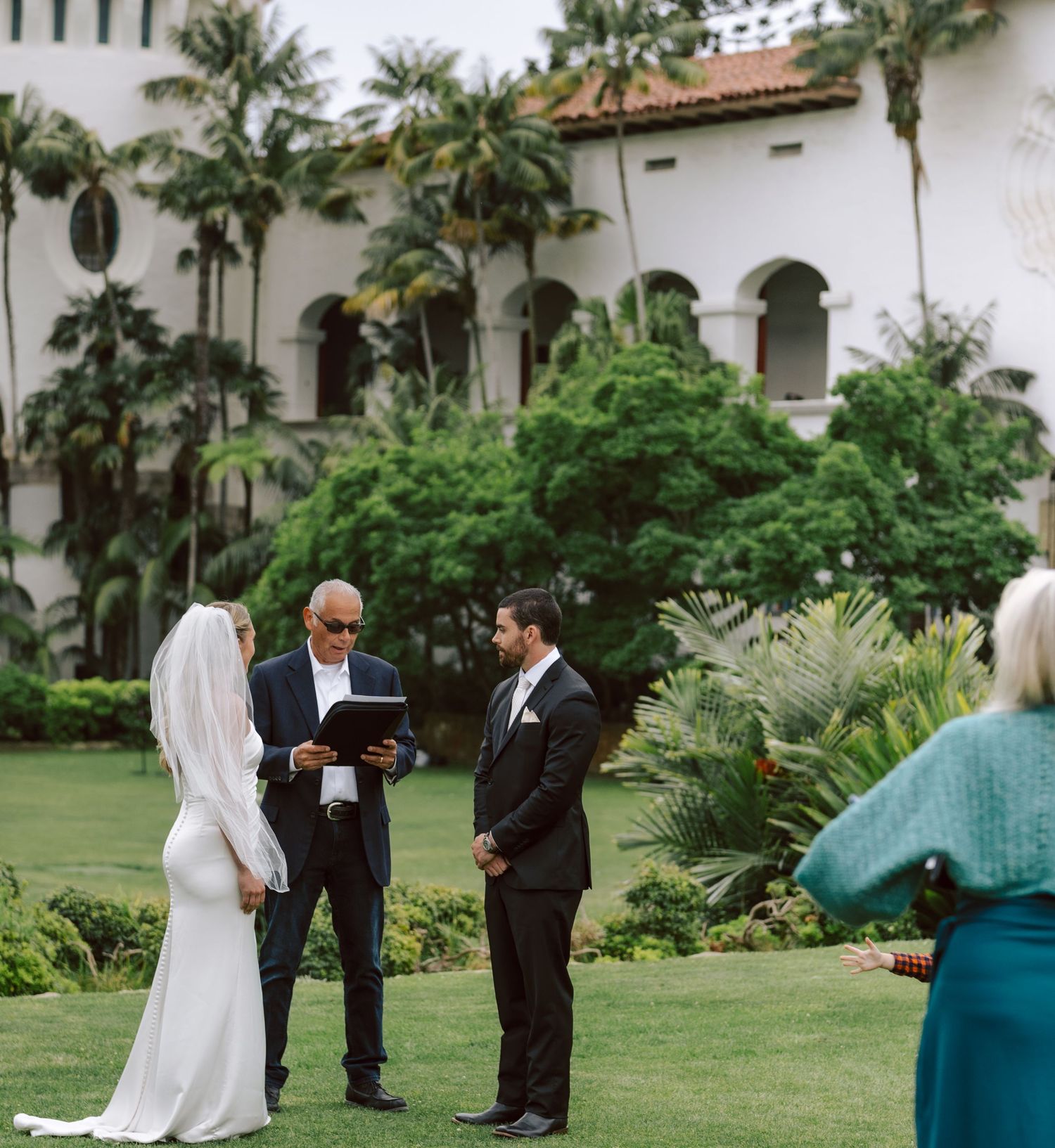 Outdoor wedding ceremony on green lawn with bride, groom, and officiant before a Spanish-style mansion.