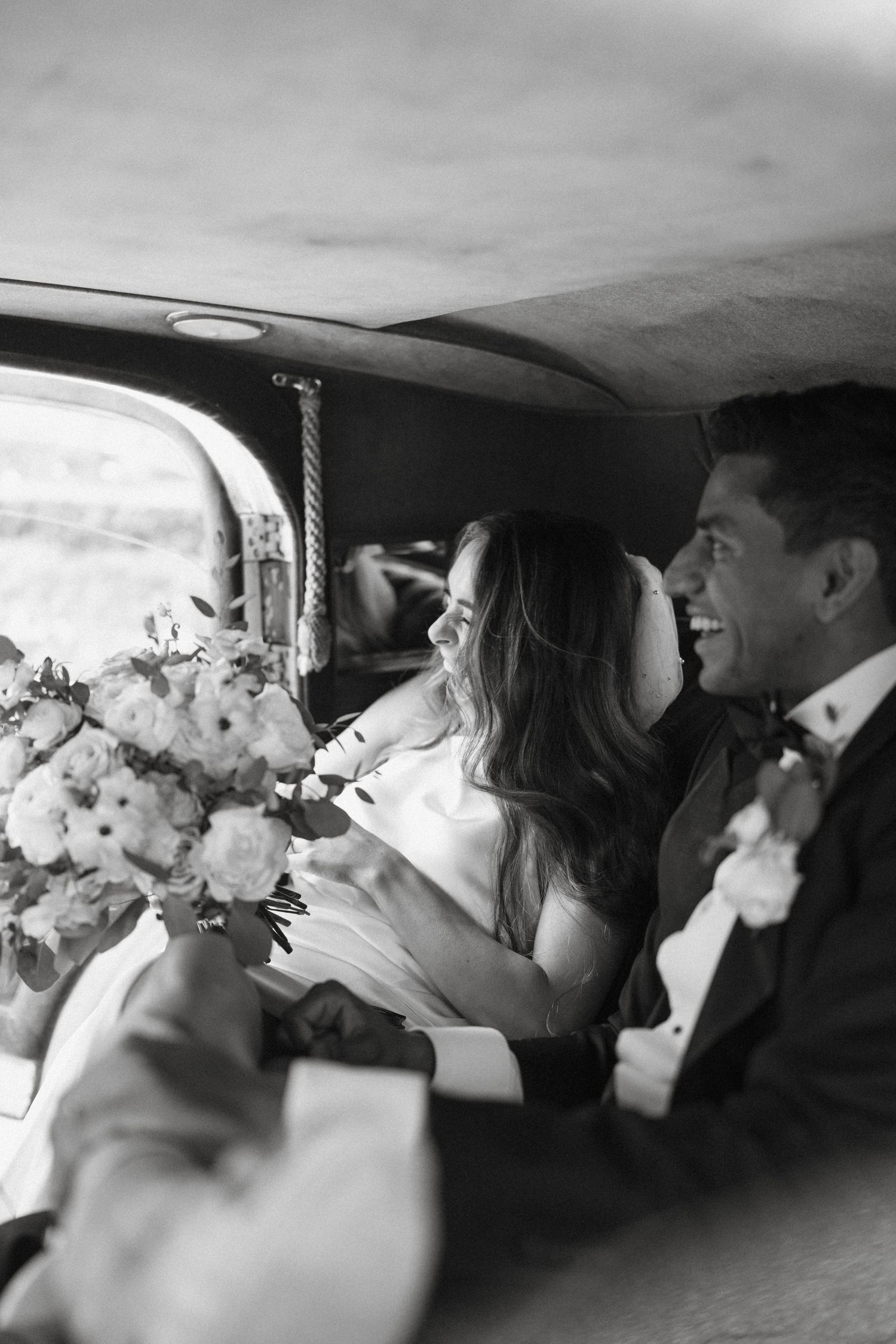 Black and white photo of a joyful bride and groom riding together in a vintage wedding car.