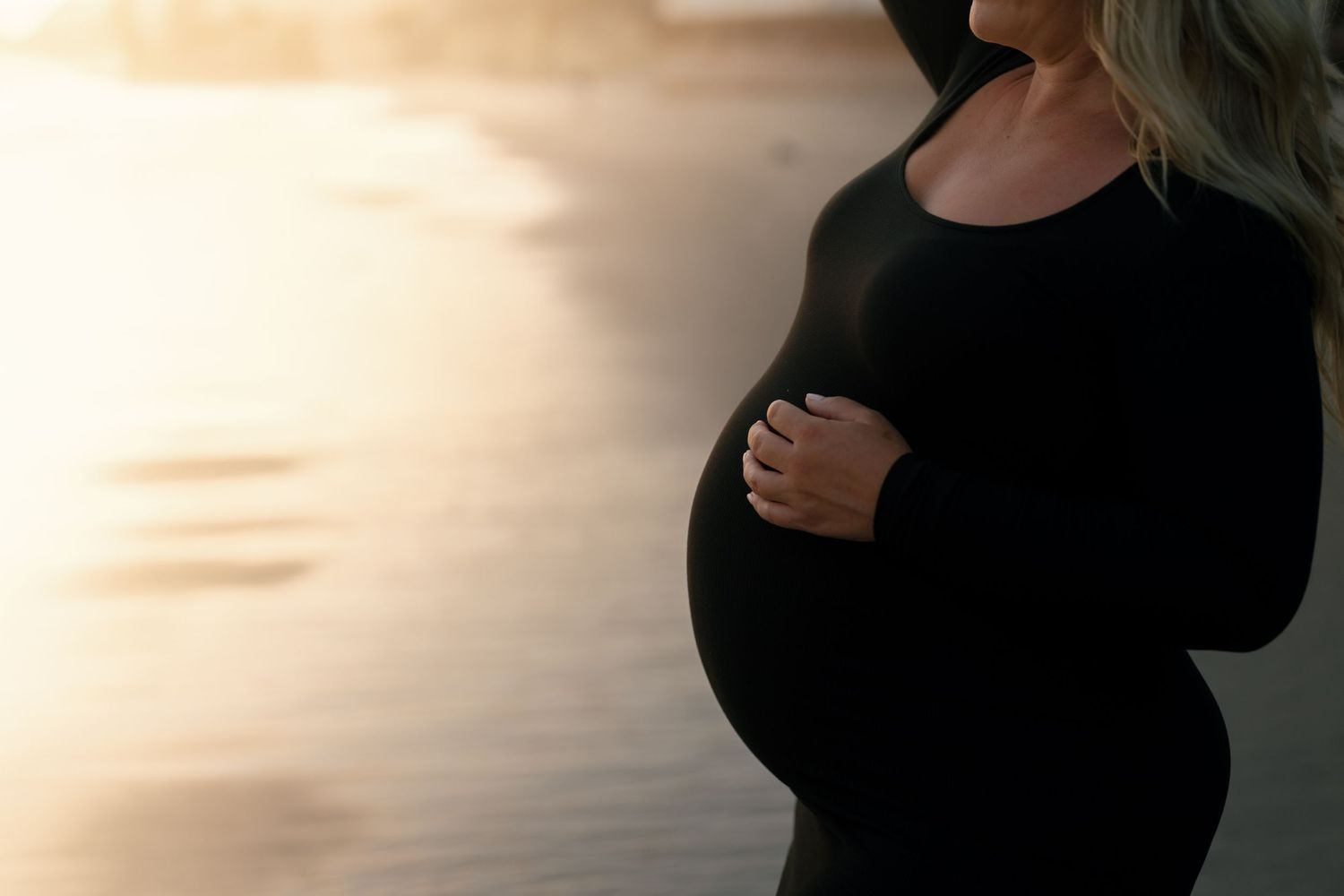Close-up of pregnant woman in black dress cradling baby bump at golden hour.