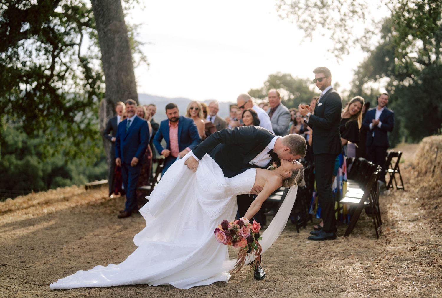 Groom dips bride in a romantic kiss after outdoor wedding ceremony surrounded by cheering guests.