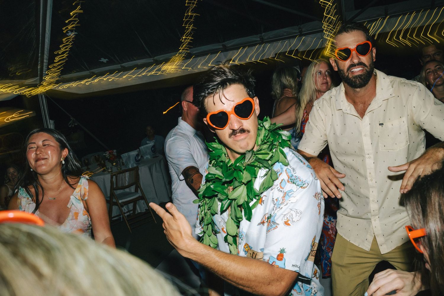 Guest in Hawaiian shirt and lei wearing novelty glasses dancing at a lively tropical wedding reception.