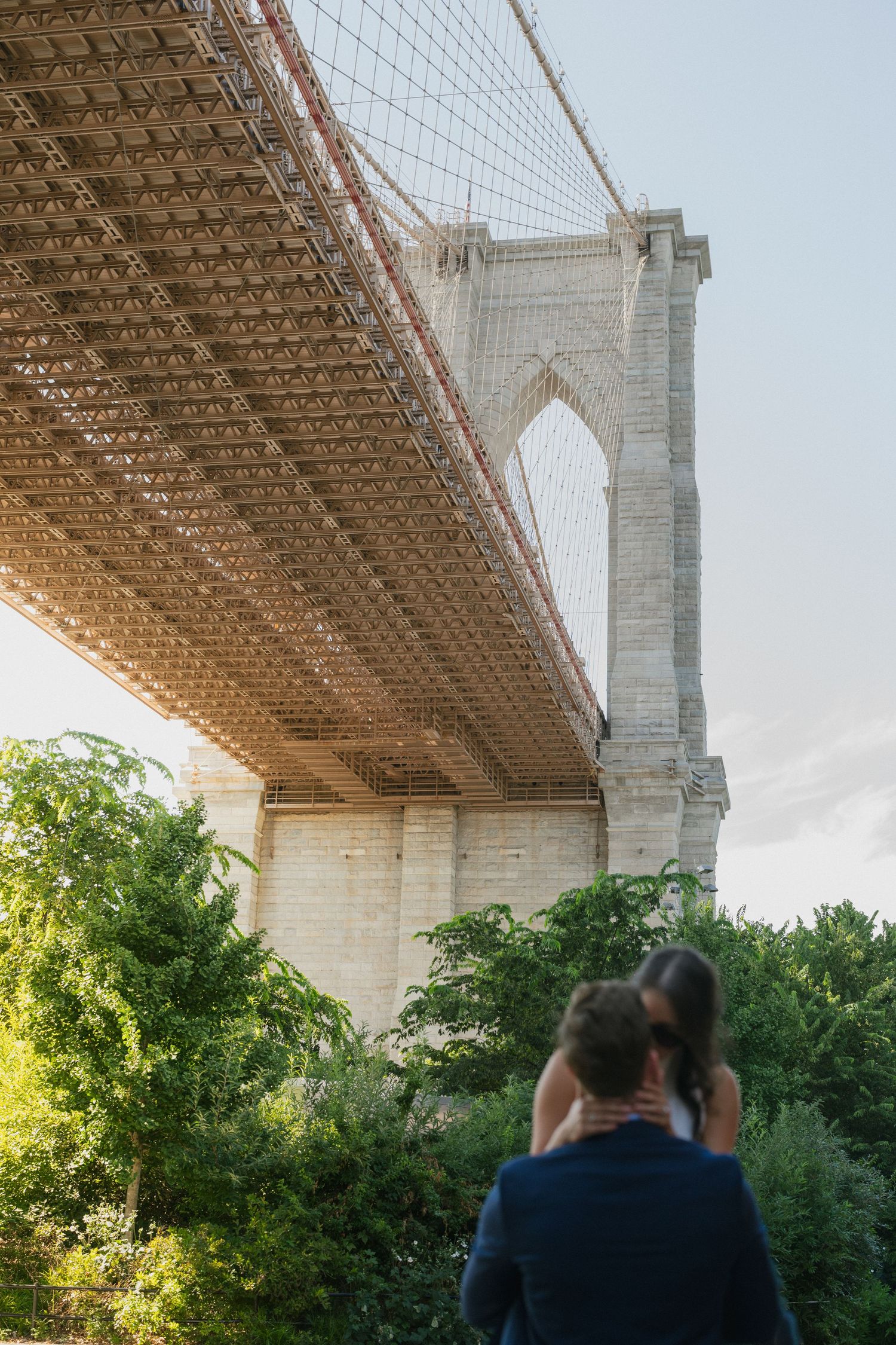 Couple embracing beneath the iconic Brooklyn Bridge surrounded by lush green trees during their engagement session.