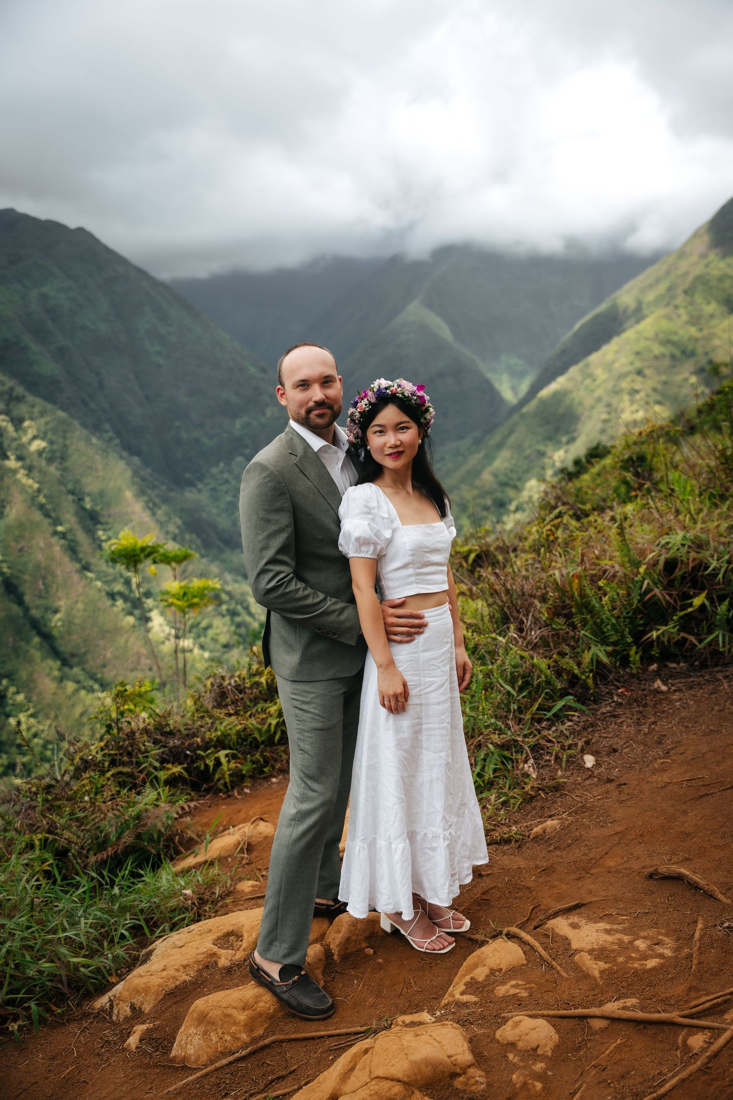 A couple poses together on a mountain trail with misty green peaks in the background for their elopment.