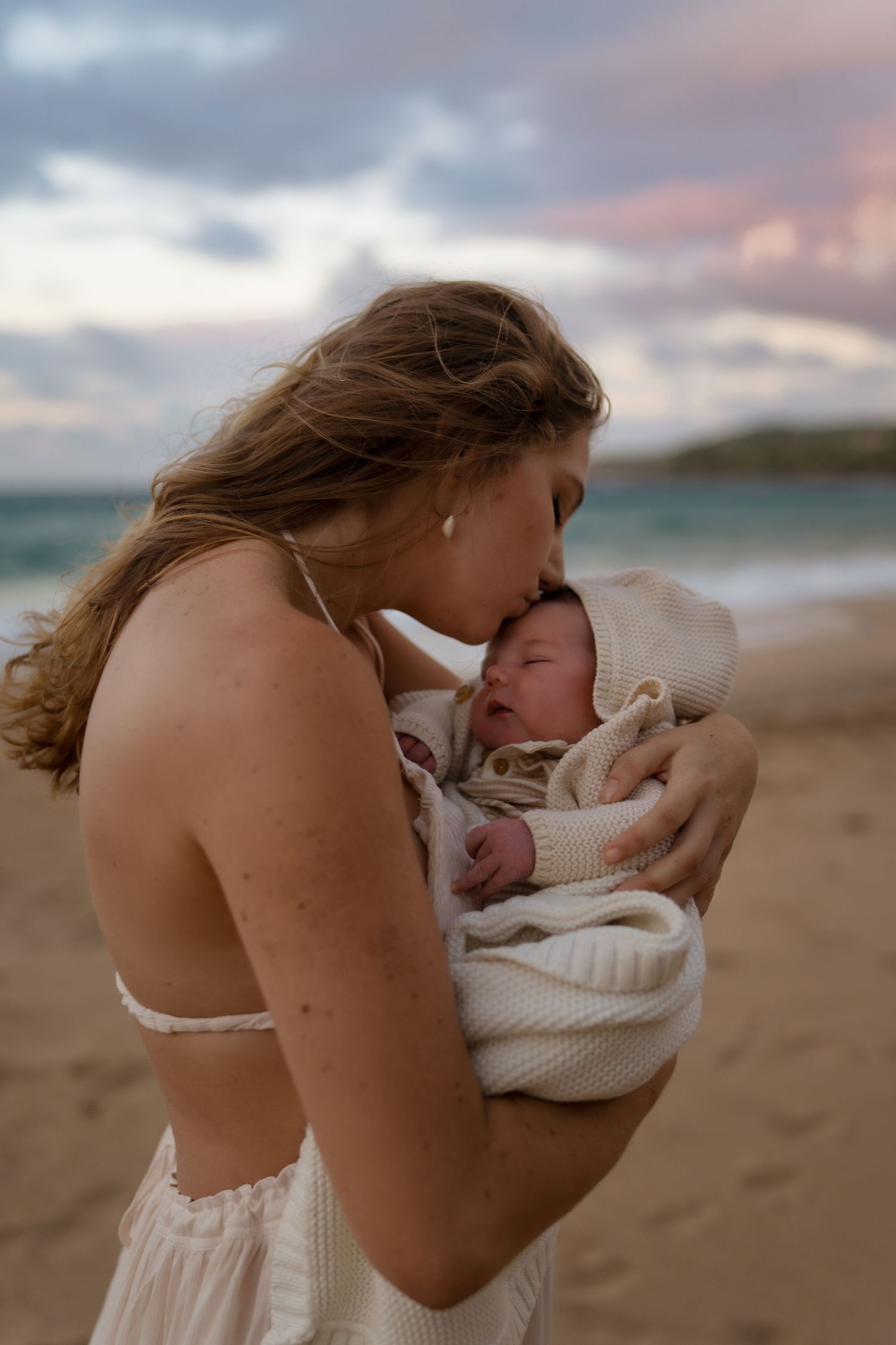 A tender moment between parent and newborn baby on a beach at sunset.