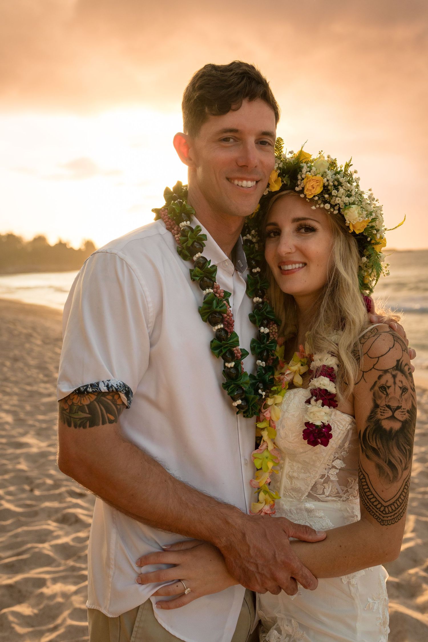 Romantic beach wedding couple embraces during golden hour sunset with floral leis and tropical decor.
