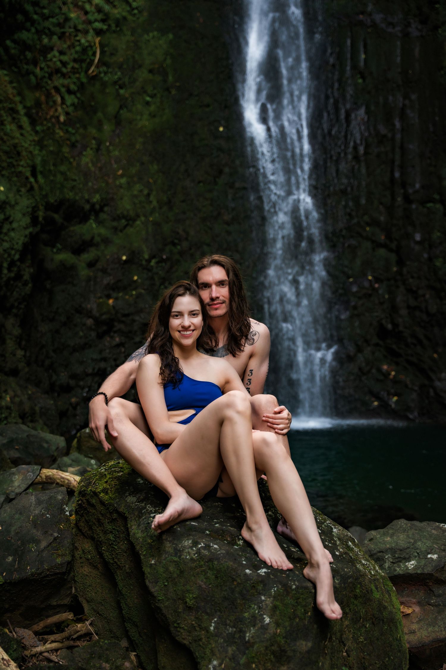 Couple poses on dark volcanic rocks with dramatic waterfall in background.
