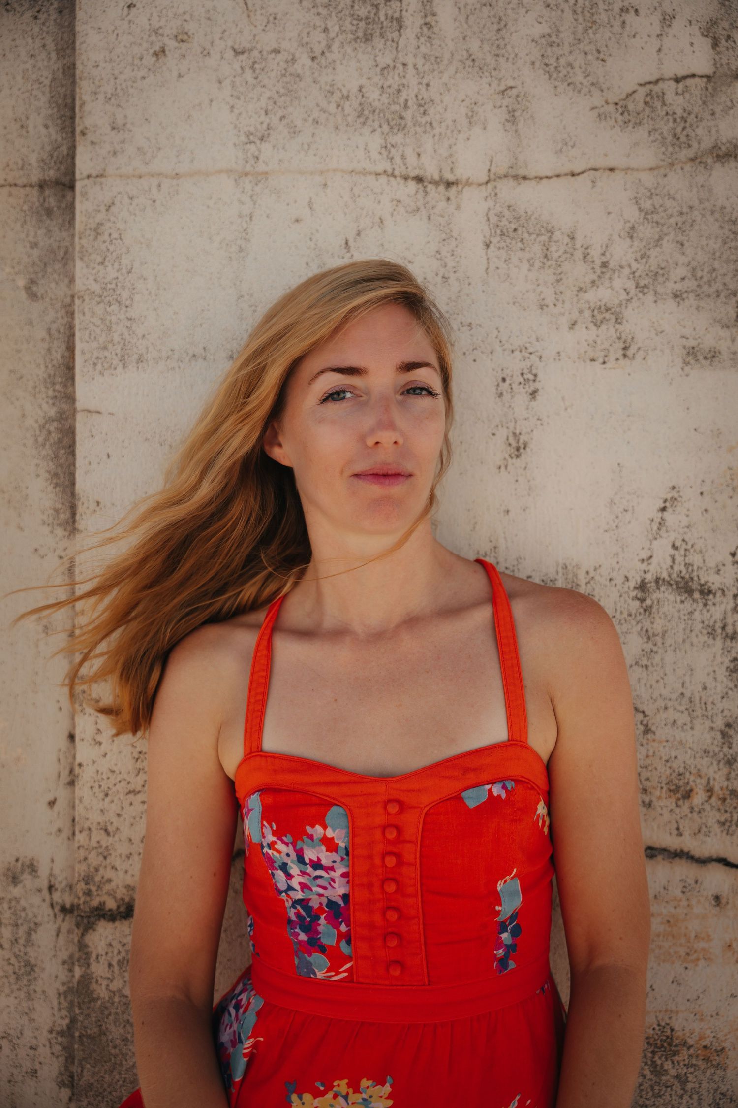 Woman in red floral dress posing against concrete wall with windswept hair.